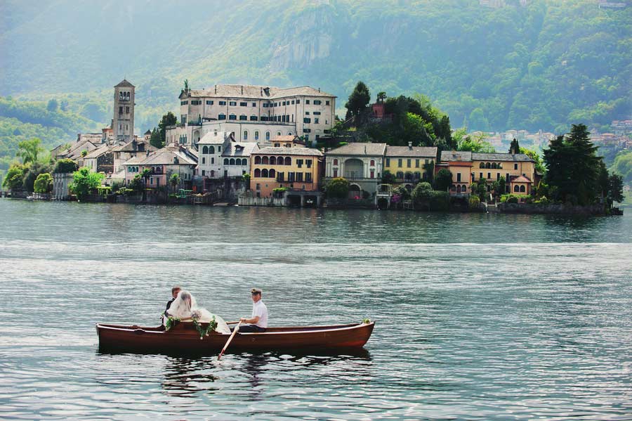 Two people in a wooden rowboat on a calm lake with a small island in the background featuring historic buildings, a church tower, and lush greenery.