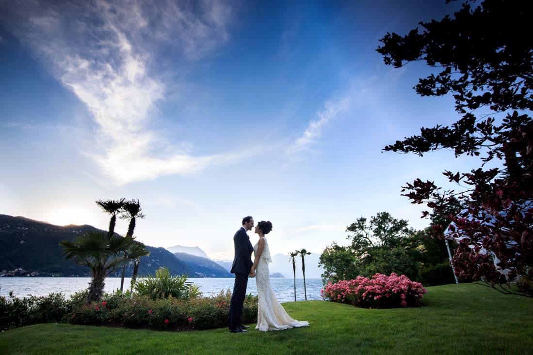 A bride and groom stand face to face on a grassy lawn by a lake, surrounded by flowers and trees, with mountains and a partly cloudy sky—an idyllic scene crafted by a skilled wedding planner in Lake Como.