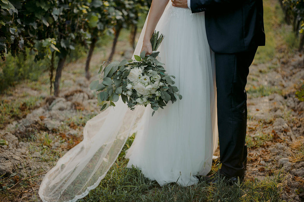 A bride in a white dress holds a bouquet of white flowers and greenery, standing next to a groom in a dark suit among grapevines. Only their lower bodies are visible. Her veil trails on the ground at this romantic Vineyard Wedding in Italy.