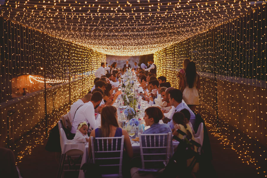 A long dining table is set outdoors under a canopy of string lights at a destination wedding in Italy, with many people seated and eating, creating a warm and festive atmosphere.
