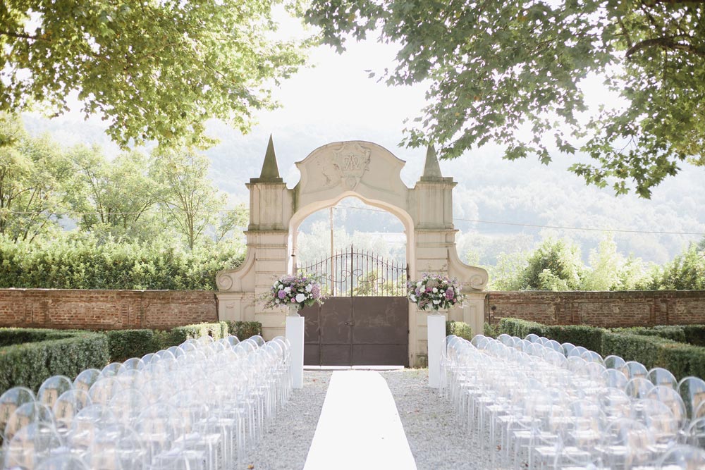 Rows of clear chairs are arranged on either side of a white aisle outdoors, leading to a decorative stone gate with two large floral arrangements. Lush greenery and trees surround the area under bright daylight.