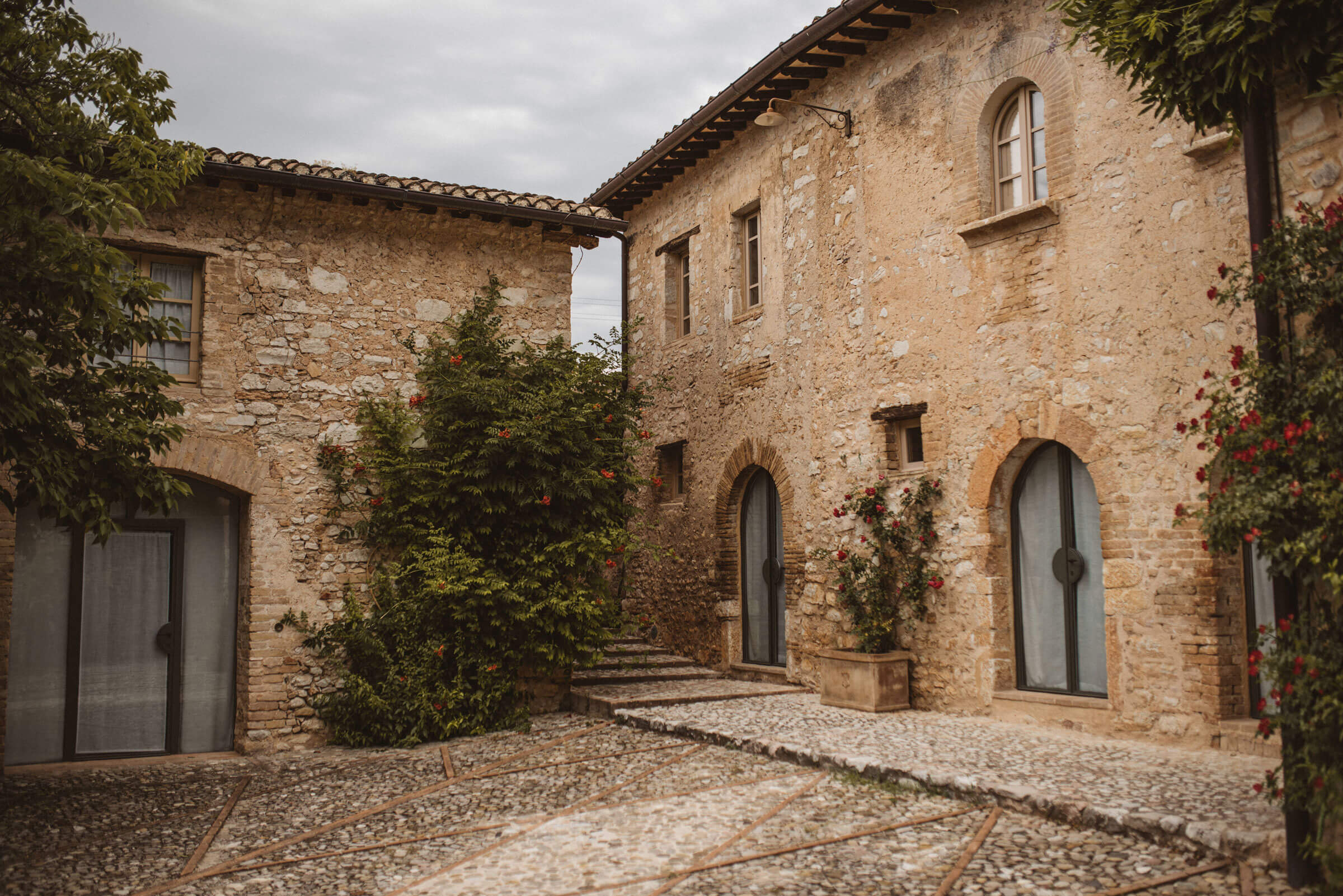 Two rustic stone buildings with arched doorways and windows stand on a cobblestone courtyard, surrounded by greenery—an idyllic setting perfect for a Destination Wedding in Umbria under a cloudy sky.