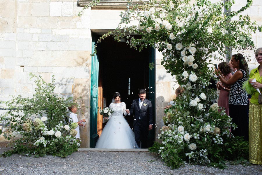 A bride in a white gown and a groom in a dark suit walk out of a stone building decorated with white flowers as people on either side watch and take photos, capturing the magic of their wedding in Tuscany.