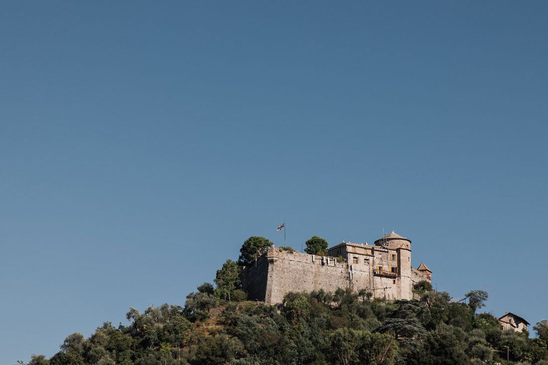 wedding-in-portofino-1 A stone fortress sits atop a tree-covered hill under a clear blue sky—a perfect backdrop for any wedding planner Liguria Italy would recommend. A flag adorns the fort, with smaller structures nestled beside the main building.
