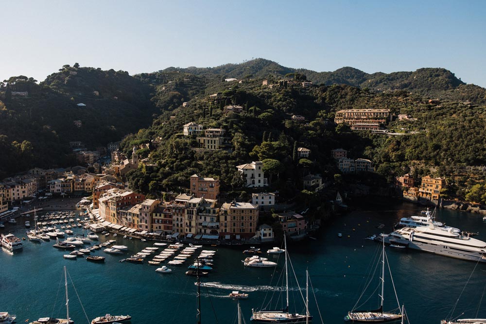 wedding-in-portofino-10 A seaside village in Liguria, Italy, with colorful buildings and boats docked in the harbor. Large yachts and green hills create a dreamy backdrop—perfect for a wedding planner seeking an unforgettable celebration by the sea.