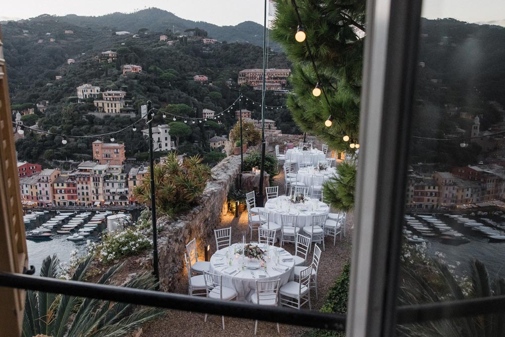 wedding-in-portofino-15 Several round tables with white tablecloths and chairs are set up outdoors on a terrace overlooking a coastal town in Liguria, Italy; string lights hang above the tables—a dream setting curated by an expert wedding planner.