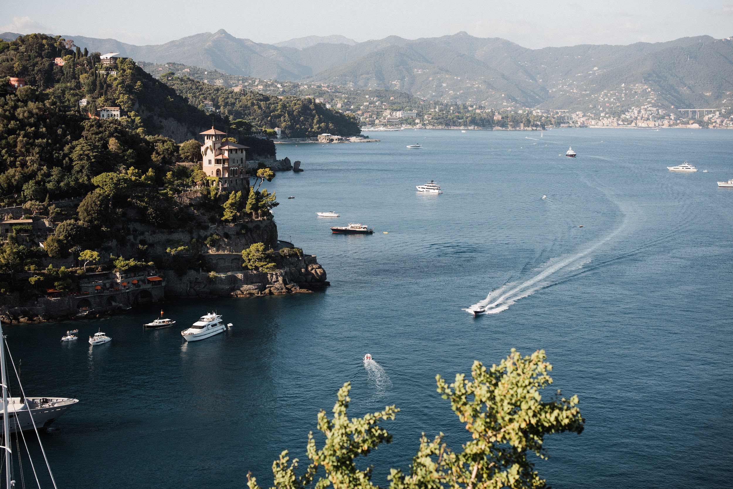 wedding-in-portofino (35) A coastal scene with several boats and yachts on blue water, a villa on a rocky forested cliff, and mountains in the background under a clear sky—an ideal setting for a wedding planner Liguria Italy could arrange. Greenery graces the foreground.