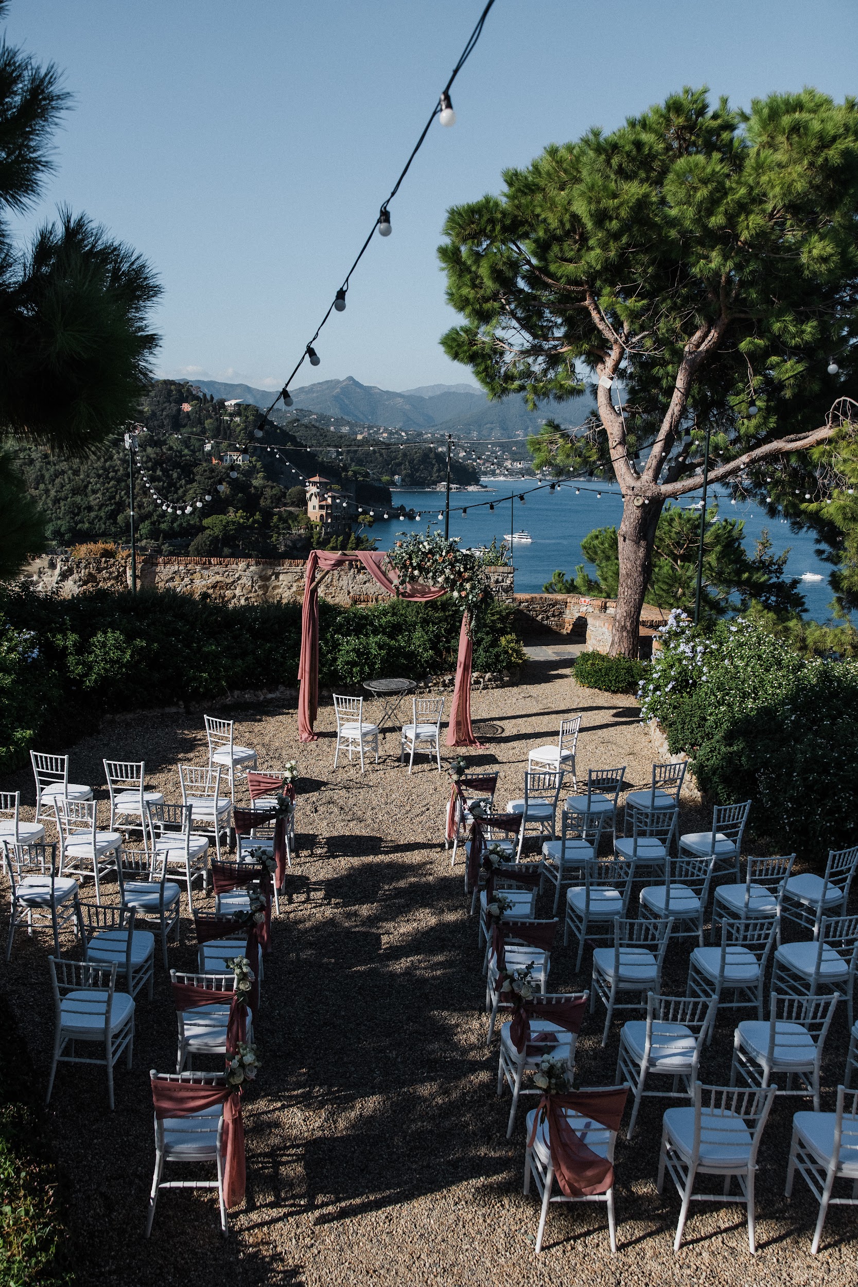 8L3A0418 Rows of white chairs line an outdoor aisle facing a wooden arch draped in fabric. String lights twinkle above, with water, hills, and trees as a backdrop—an idyllic setting crafted by a wedding planner in Liguria, Italy.