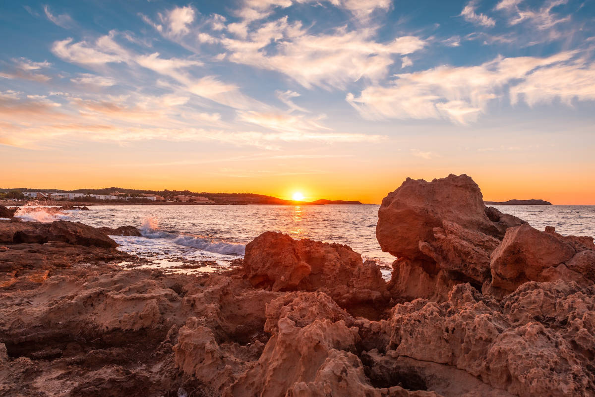 ibiza_wedding_2 Rocky shoreline at sunset, sun low on the horizon and scattered clouds in an orange-blue sky—an idyllic scene perfect for a luxury wedding planner Ibiza dreaming of stunning seaside celebrations. Calm waves and distant land complete the beautiful backdrop.