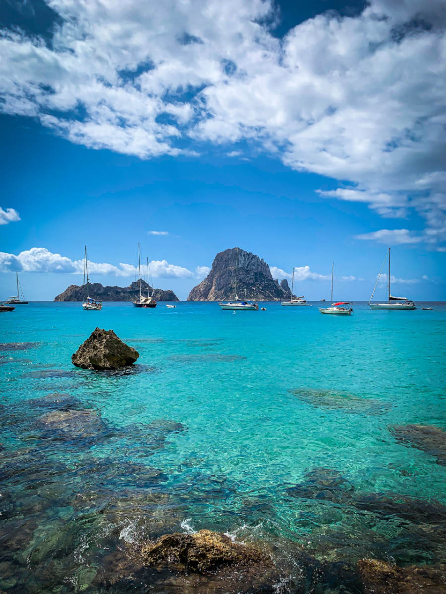 ibiza_wedding_3 Clear turquoise water with rocks in the foreground, several sailboats floating on the sea, and a large rocky island in the background—an idyllic scene perfect for a luxury wedding planner Ibiza experience under a partly cloudy sky.