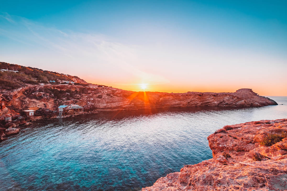 ibiza_wedding_planner The sun sets over a rocky coastline with clear blue water. With sparse vegetation on the cliffs and small buildings near the shore, it’s an idyllic scene—perfect inspiration for a luxury wedding planner Ibiza could offer.