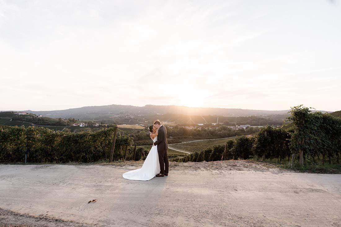 A bride and groom stand embracing on a dirt road, surrounded by vineyards in the Langhe, one of the top-wedding-venues-in-piedmont-vineyards, with rolling hills and a setting sun in the background.