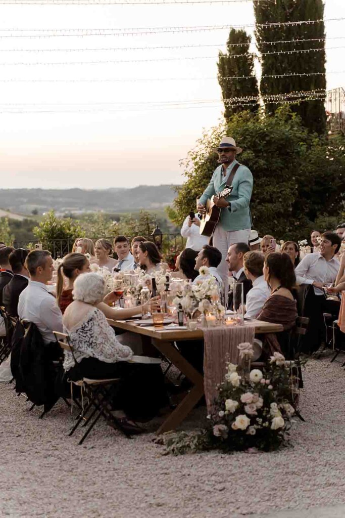 At a destination wedding in Villa Mombaruzzo, guests sit at outdoor wooden tables adorned with flowers as a musician in a light blue jacket and hat plays guitar and sings on stage at sunset, with string lights glowing above the celebration.