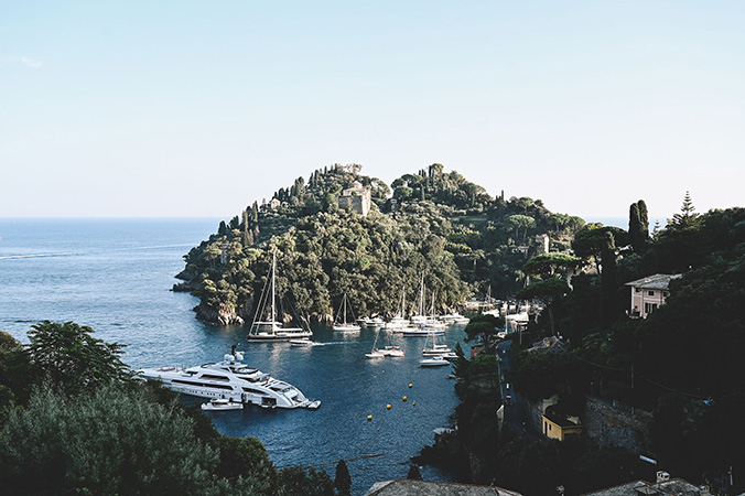 A small romantic harbor in Portofino with white yachts and sailboats docked, surrounded by lush green hills and Mediterranean-style buildings, offering a stunning sea view under a clear blue sky—perfect for a wedding.