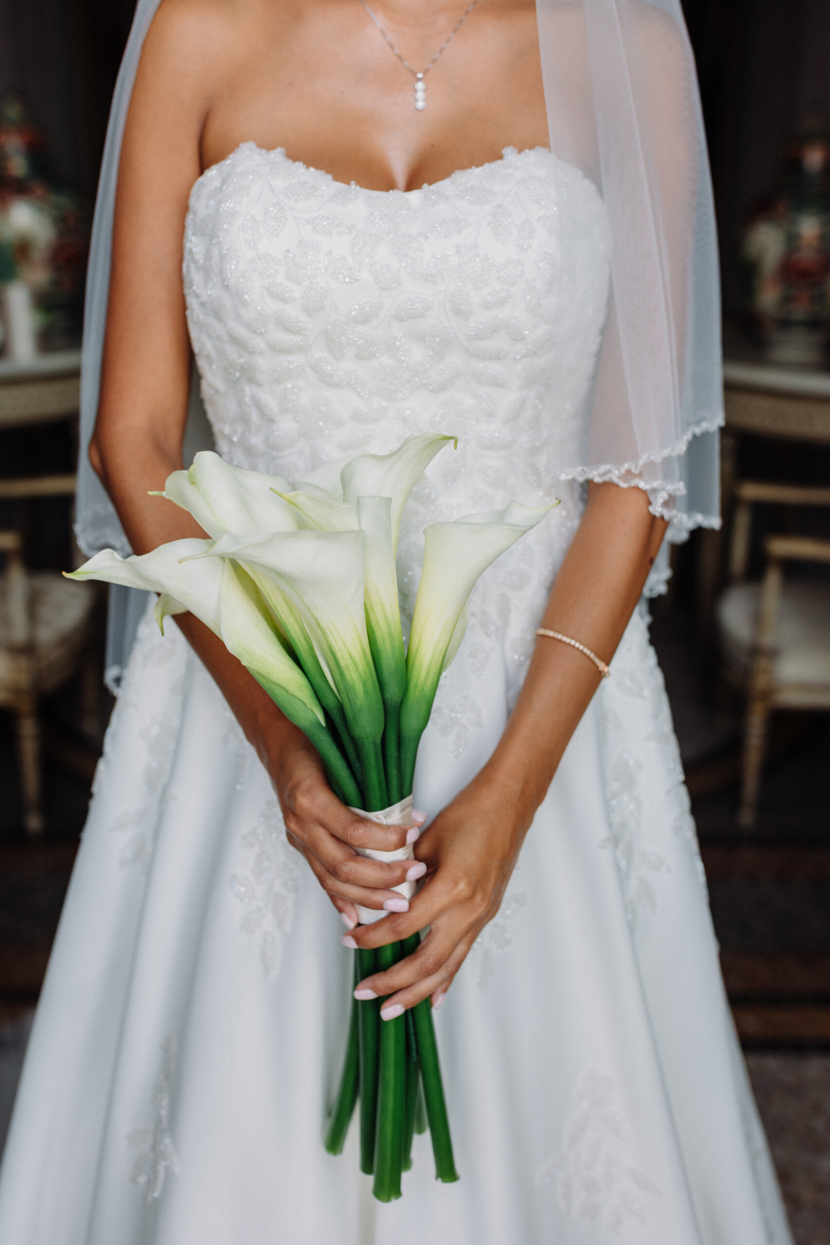 White Calla Lily Bridal Bouquet – Villa Cimena Wedding Elegant bridal bouquet of white calla lilies with long green stems, held against a white wedding dress with floral embroidery.
