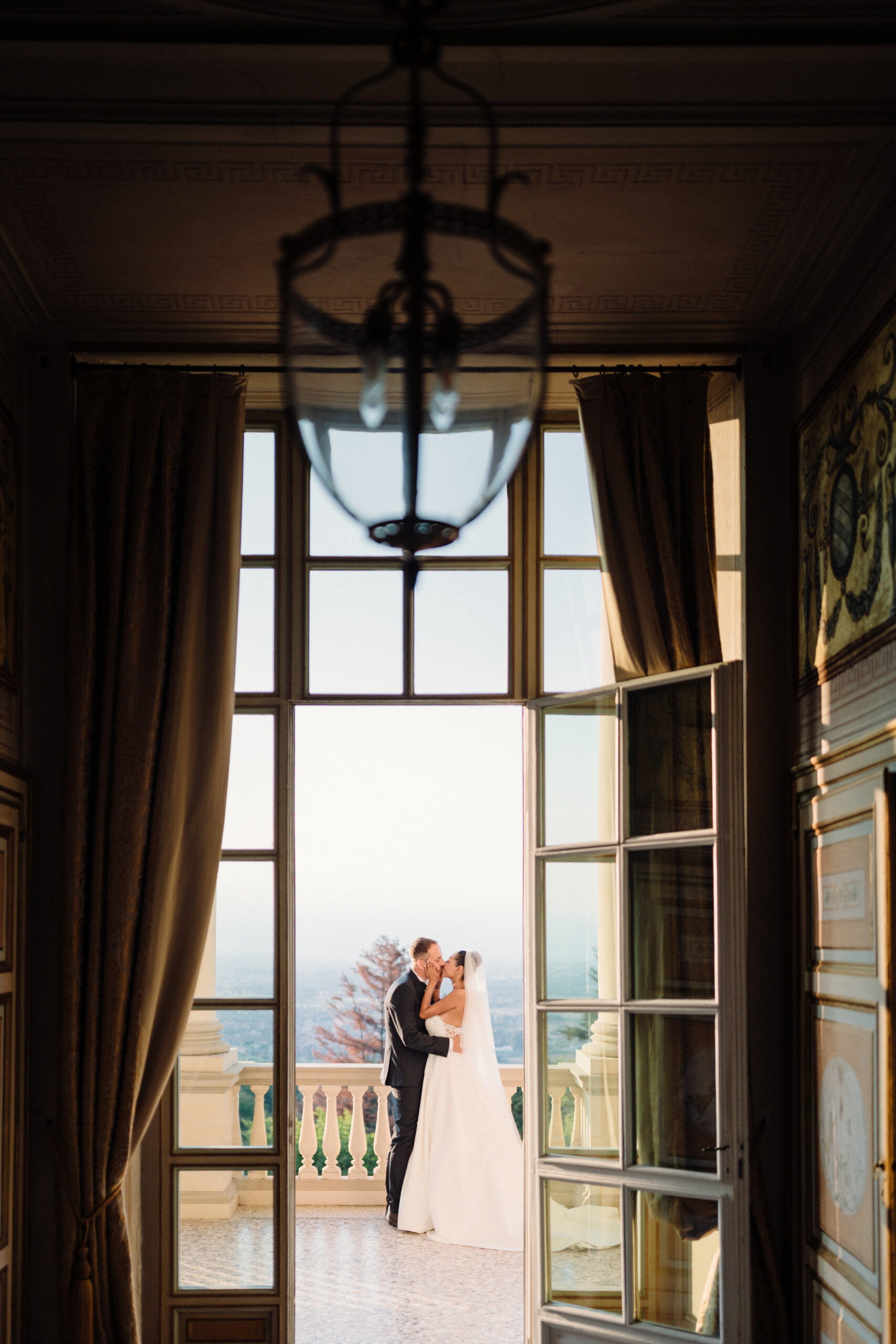 Romantic Balcony Kiss – Villa Cimena Wedding in Italy Giannella and Enrico kissing on the balcony of Villa Cimena, framed by a large window and elegant historic interiors.