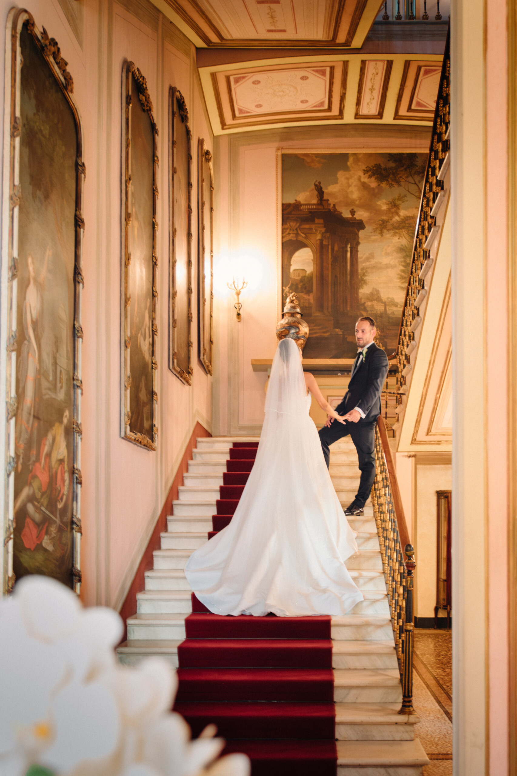 Giannella and Enrico holding hands on the grand red-carpeted staircase of Villa Cimena, surrounded by historic paintings and ornate interiors.