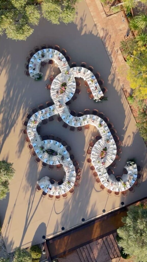 Overhead view of white serpentine wedding tables with floral centrepieces and round chairs arranged on a green lawn.